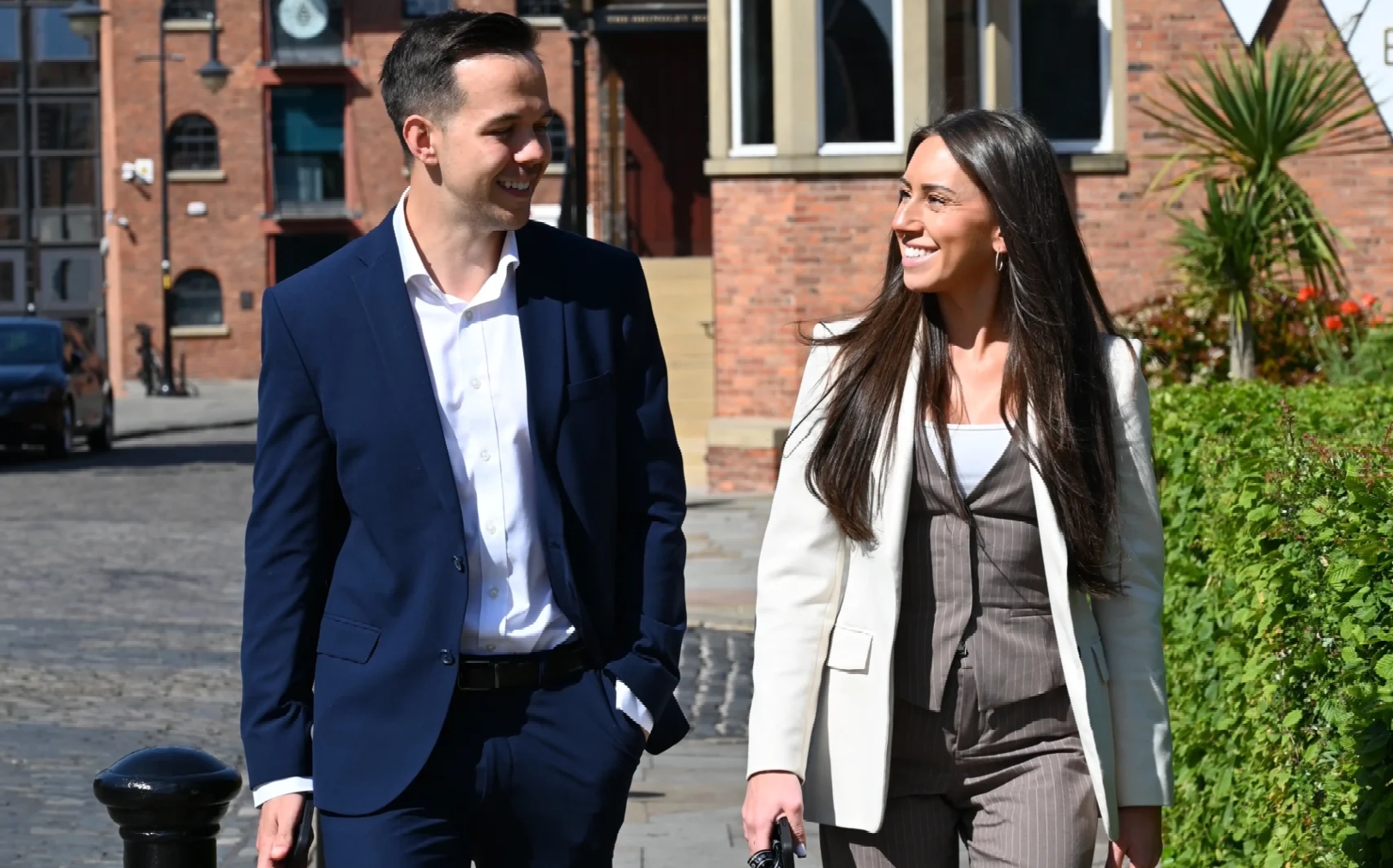 A man and woman both wearing business suits walking down a road near an office building